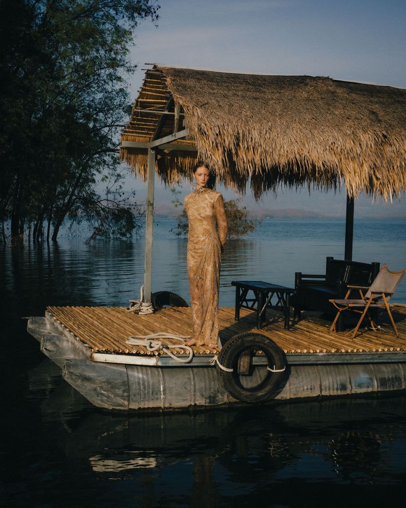 Model Lydia Reid poses in a floor-length patterned beige dress on a thatched bamboo floating platform over calm lake water.