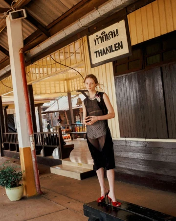 Model Lydia Reid standing on a wooden bench under a train station sign reading Thakilen in a sheer black outfit and red heels.