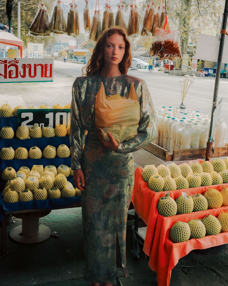 A woman in a metallic floral dress holding a plastic bag stands at a street market stall filled with netting-wrapped fruits.
