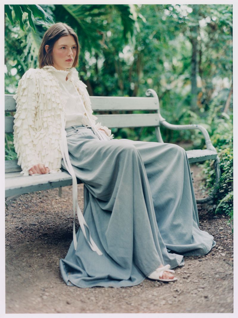 Model Anna Viksoe wearing a white fringe jacket and voluminous slate blue trousers seated on a garden bench surrounded by lush greenery.