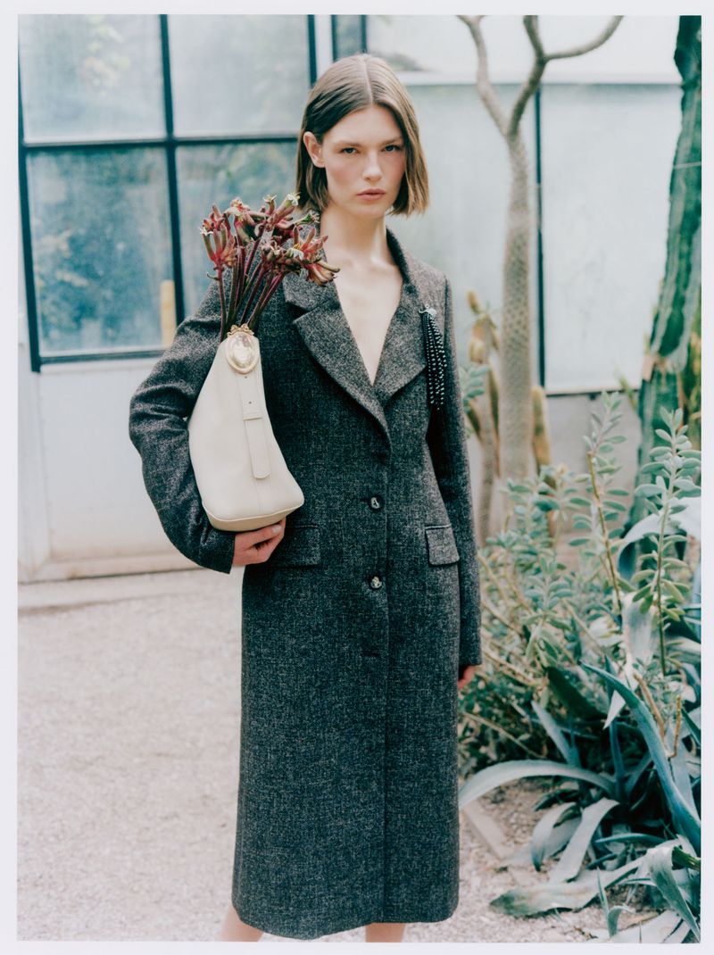 Model Anna Viksoe in a long charcoal tweed coat holding a cream handbag filled with exotic plants in a greenhouse.