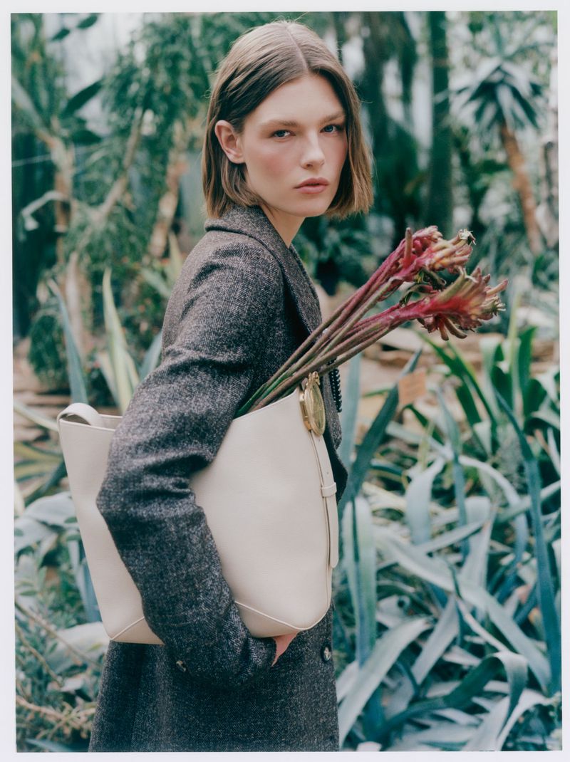 Model Anna Viksoe wearing a tweed coat and holding a cream leather tote bag filled with red botanical stems in a garden.