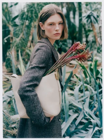 Model Anna Viksoe wearing a tweed coat and holding a cream leather tote bag filled with red botanical stems in a garden.