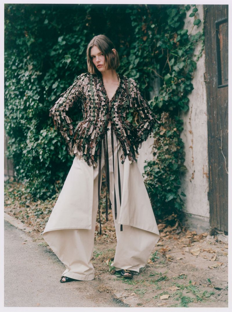 Model Anna Viksoe wearing a textured tassel jacket and sculptural wide-leg cream trousers against an ivy-covered rustic wall.
