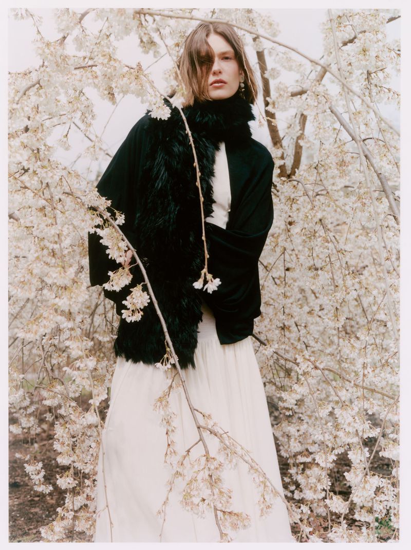 Model Anna Viksoe standing amidst blooming white blossom branches wearing a black jacket with a fur collar and a long white skirt.