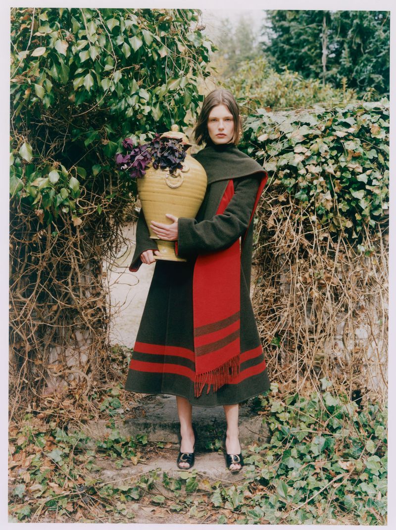 Model Anna Viksoe in an olive green coat holding a large yellow ceramic urn amidst overgrown ivy in a garden.