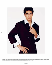 Editorial portrait of a man posing in a dark tailored double-breasted blazer and open-collar white shirt against a white backdrop.