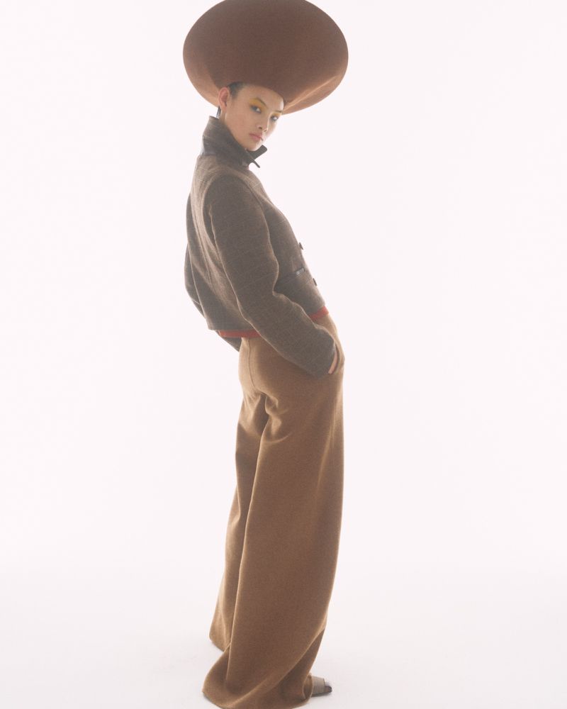 Model wearing an oversized sculptural brown dome hat, cropped jacket, and wide-leg brown trousers against a bright white background.