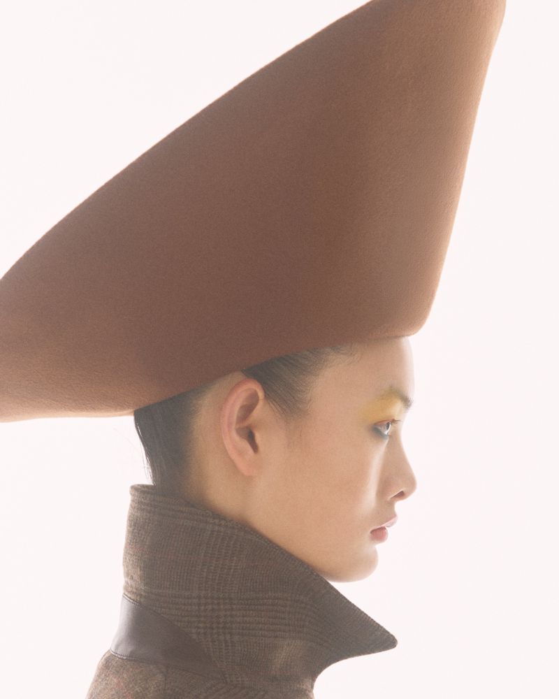 Side profile of an East Asian woman wearing an oversized conical brown felt hat and a high-collar coat against a white background.