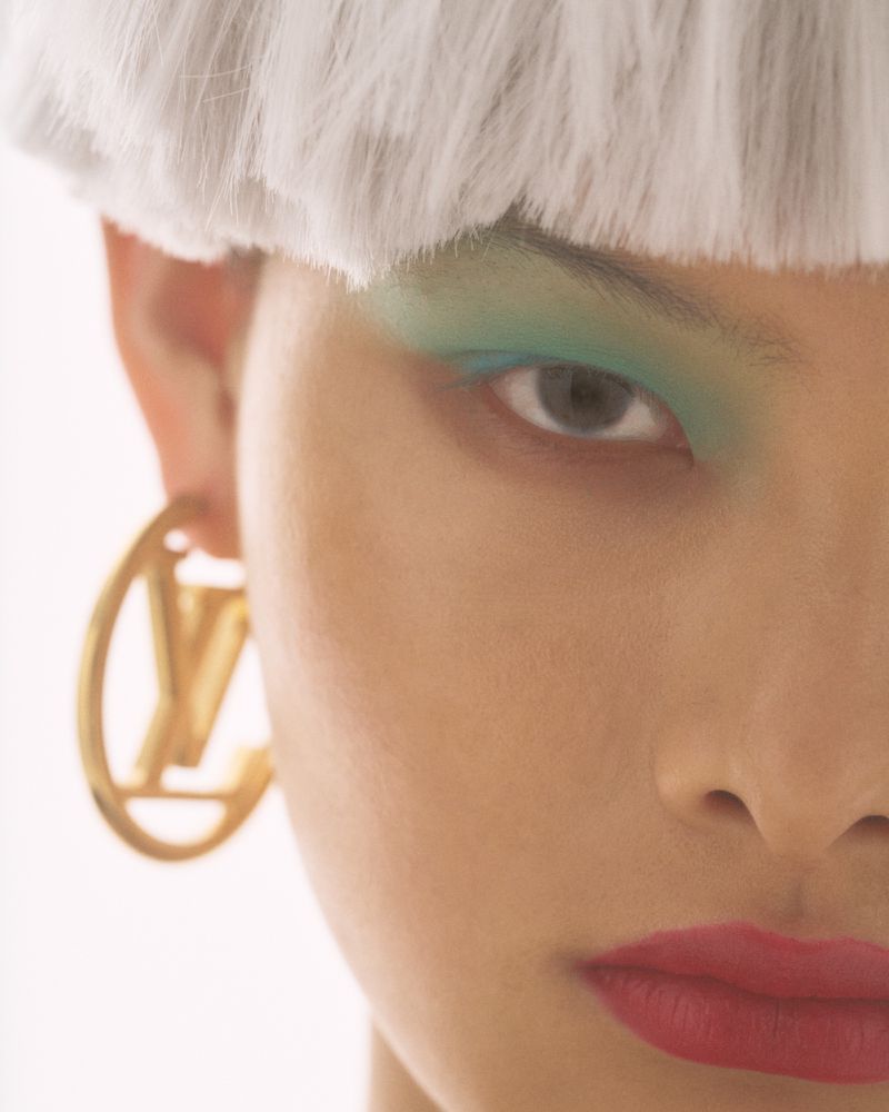 Close-up portrait of a model with turquoise eyeshadow, red lips, gold hoop earrings, and a white fringed hairpiece for Inflate Deflate.