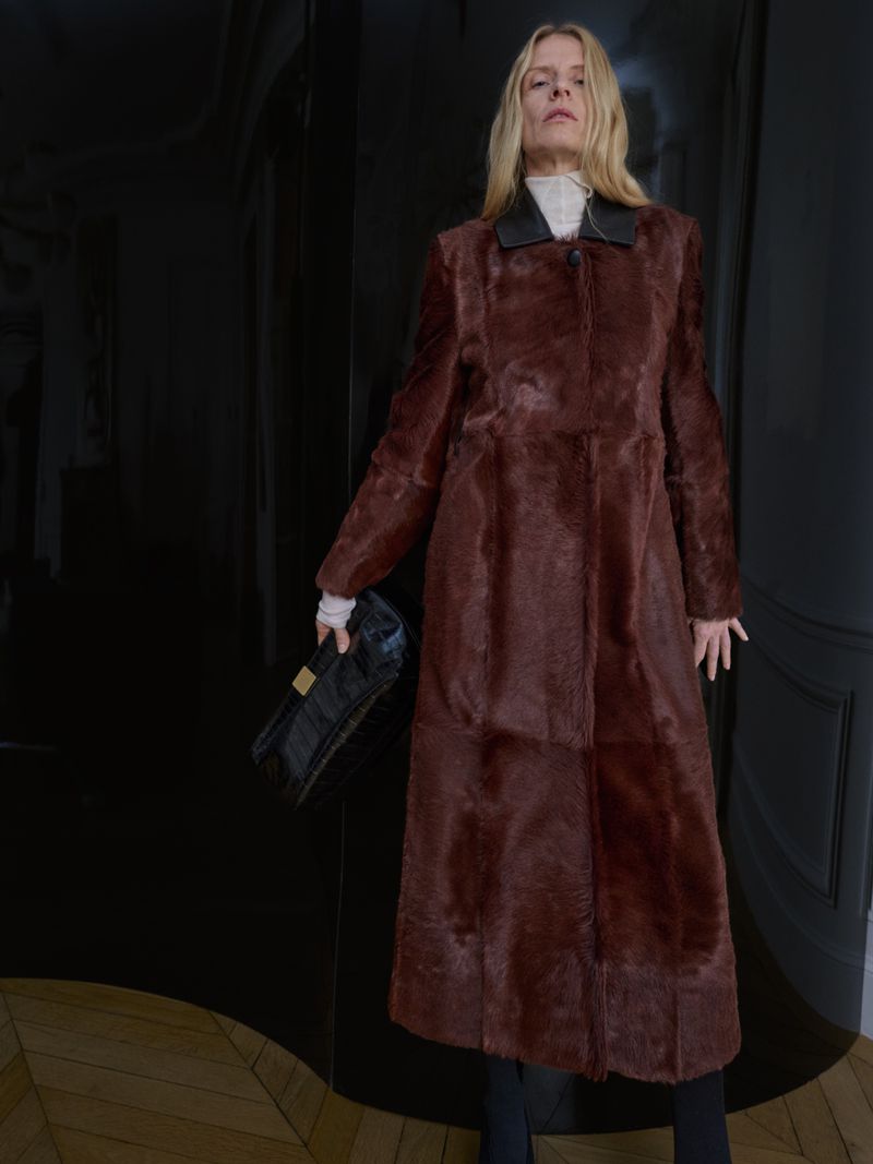 Model wearing a full-length rich brown textured fur coat with a black collar and holding a black clutch bag.