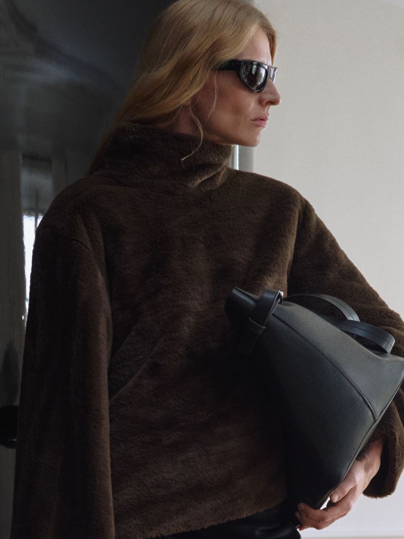 A woman wearing dark sunglasses and a brown textured high-neck top while holding a structured black leather tote bag under her arm.
