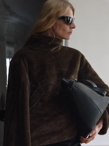 A woman wearing dark sunglasses and a brown textured high-neck top while holding a structured black leather tote bag under her arm.