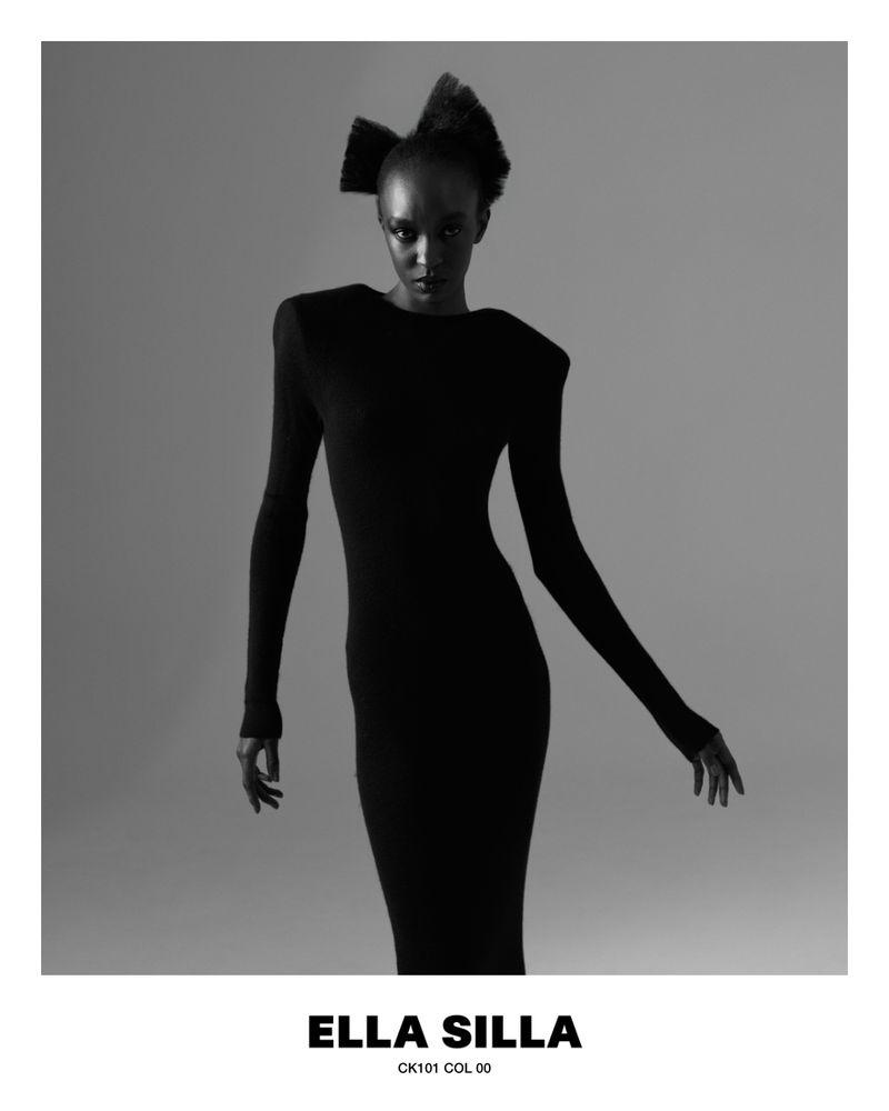 Model Nicole Atieno wears a fitted black long-sleeve dress with structured shoulders against a plain grey studio backdrop.