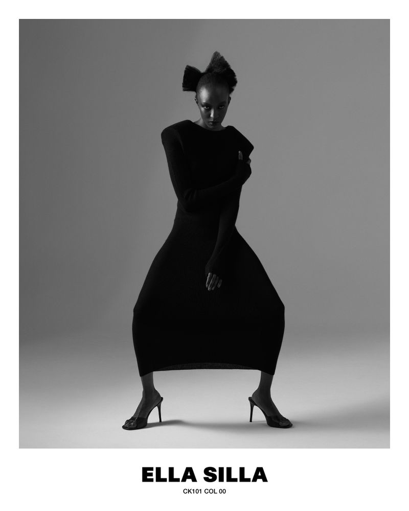 Nicole Atieno posing in a black long-sleeve dress and stiletto heels against a grey seamless background for Ella Silla.