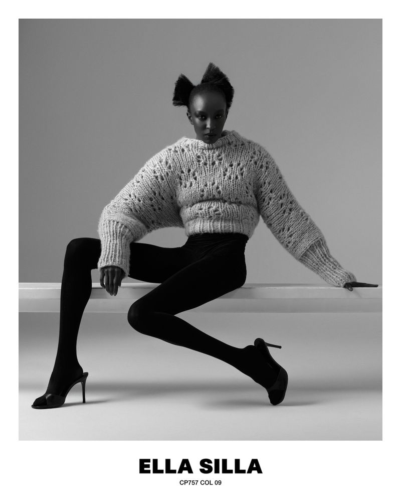 Nicole Atieno seated on a white bench wearing an oversized chunky knit sweater, black tights, and high heels in black and white.
