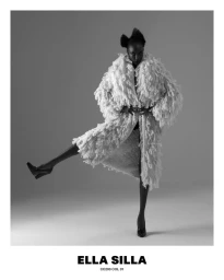 Model Nicole Atieno in a textured fringed coat and heels posing with one leg raised against a studio backdrop.