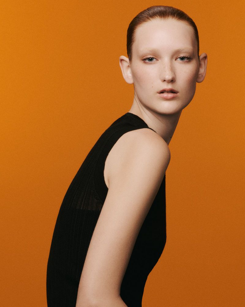 Studio portrait of a fair-skinned woman with slicked-back auburn hair wearing a black tank top against an orange backdrop.