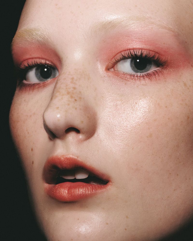 Close-up studio shot of a fair-skinned young adult female with pink eye shadow, coral lipstick, and freckles against a dark backdrop.