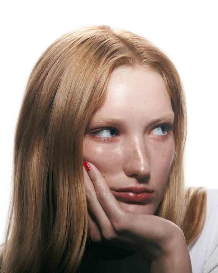 Close-up portrait of a model with reddish-blonde hair resting her hand against her cheek with red nail polish
