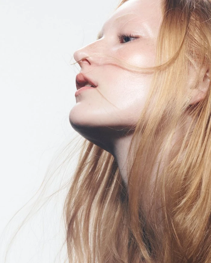 Close-up profile portrait of a model with blonde hair and glowing skin against a plain white background.