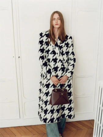 Model in a black and white houndstooth coat holding a dark brown handbag standing against panelled doors