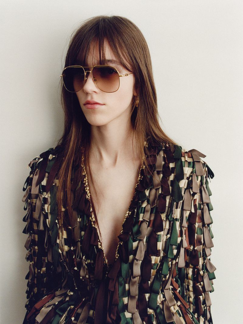 Model wearing gold-tone aviator sunglasses and a multicolored ribbon-fringe jacket against an off-white background.