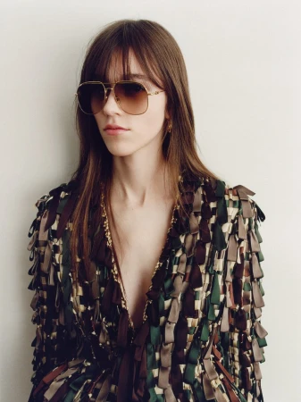 Model wearing gold-tone aviator sunglasses and a multicolored ribbon-fringe jacket against an off-white background.