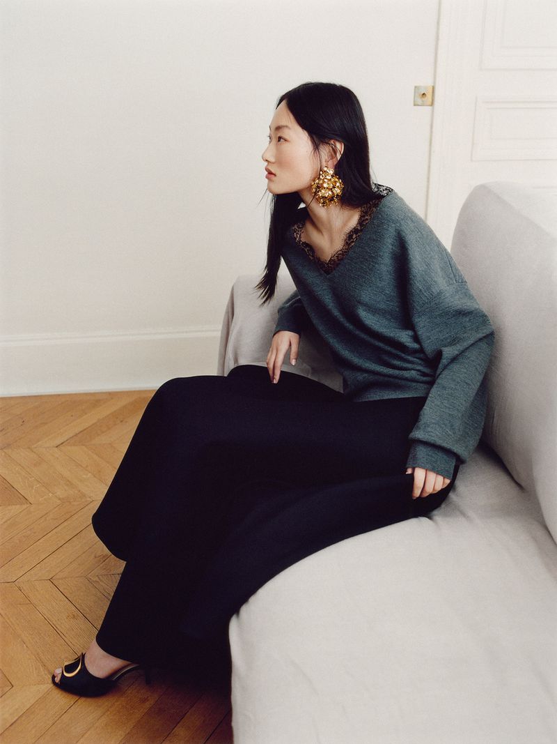 Model seated on a light grey sofa wearing a grey long-sleeve top, black skirt, and large gold statement earrings.