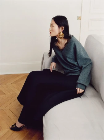 Model seated on a light grey sofa wearing a grey long-sleeve top, black skirt, and large gold statement earrings.