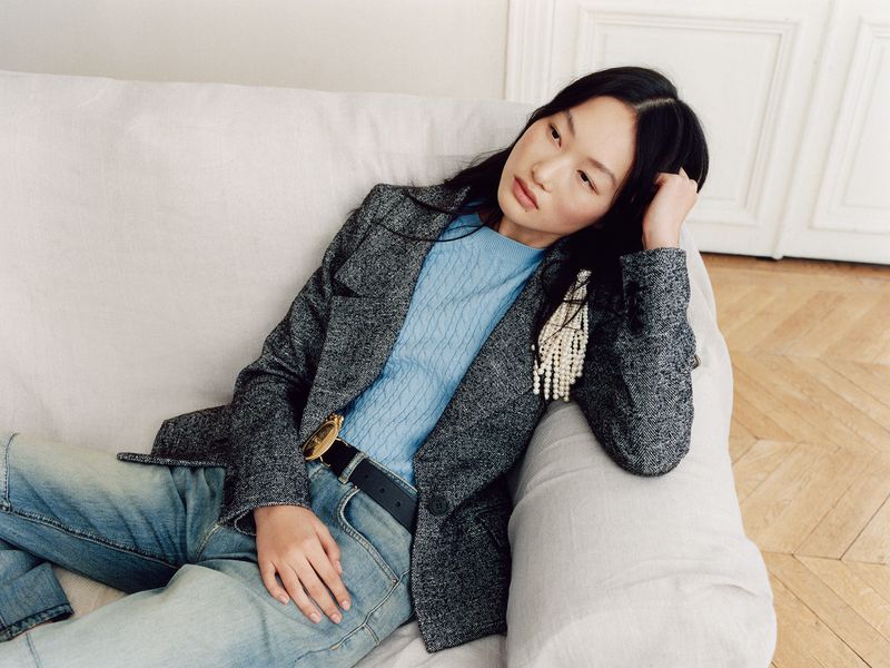 An East Asian woman reclines on a white sofa wearing a grey tweed blazer, light blue knit top, and faded denim jeans.