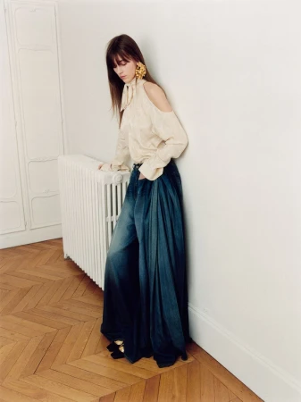 Model leaning against a radiator wearing a cream cold-shoulder blouse and wide denim trousers on parquet flooring.