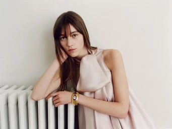 A model in a pale blush satin high-neck dress leaning on a white radiator and wearing a wide gold cuff watch.