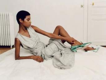 Model in a draped silver-grey sleeveless dress and metallic green kitten heels reclining on a white floor sheet against a white wall.