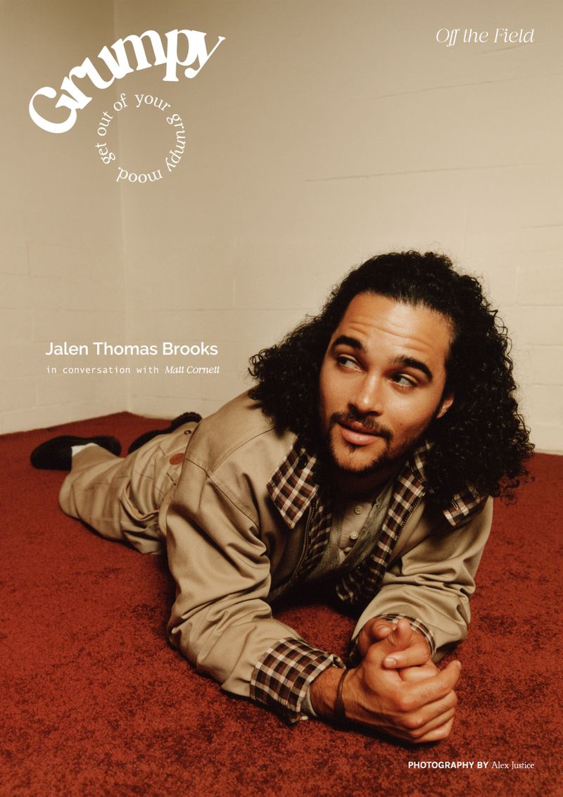 Jalen Thomas Brooks lying on a reddish-brown carpet wearing a tan jacket with plaid trim against plain white walls.