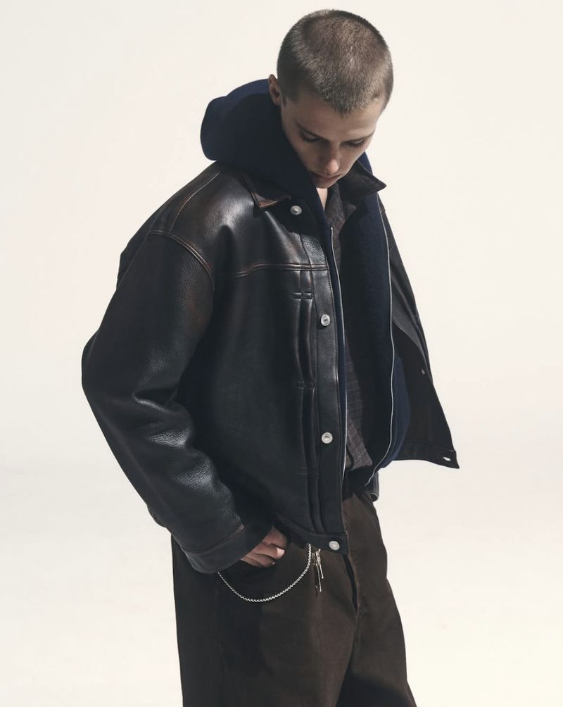 Model Elias Monstrey wearing an oversized black leather jacket and dark hoodie against a white studio background.