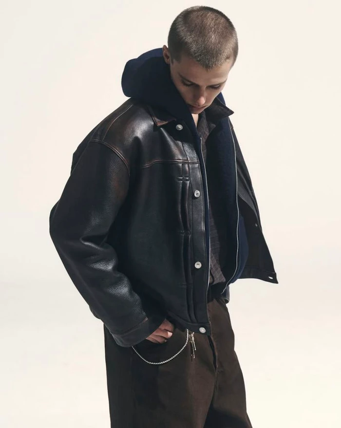 Model Elias Monstrey wearing an oversized black leather jacket and dark hoodie against a white studio background.