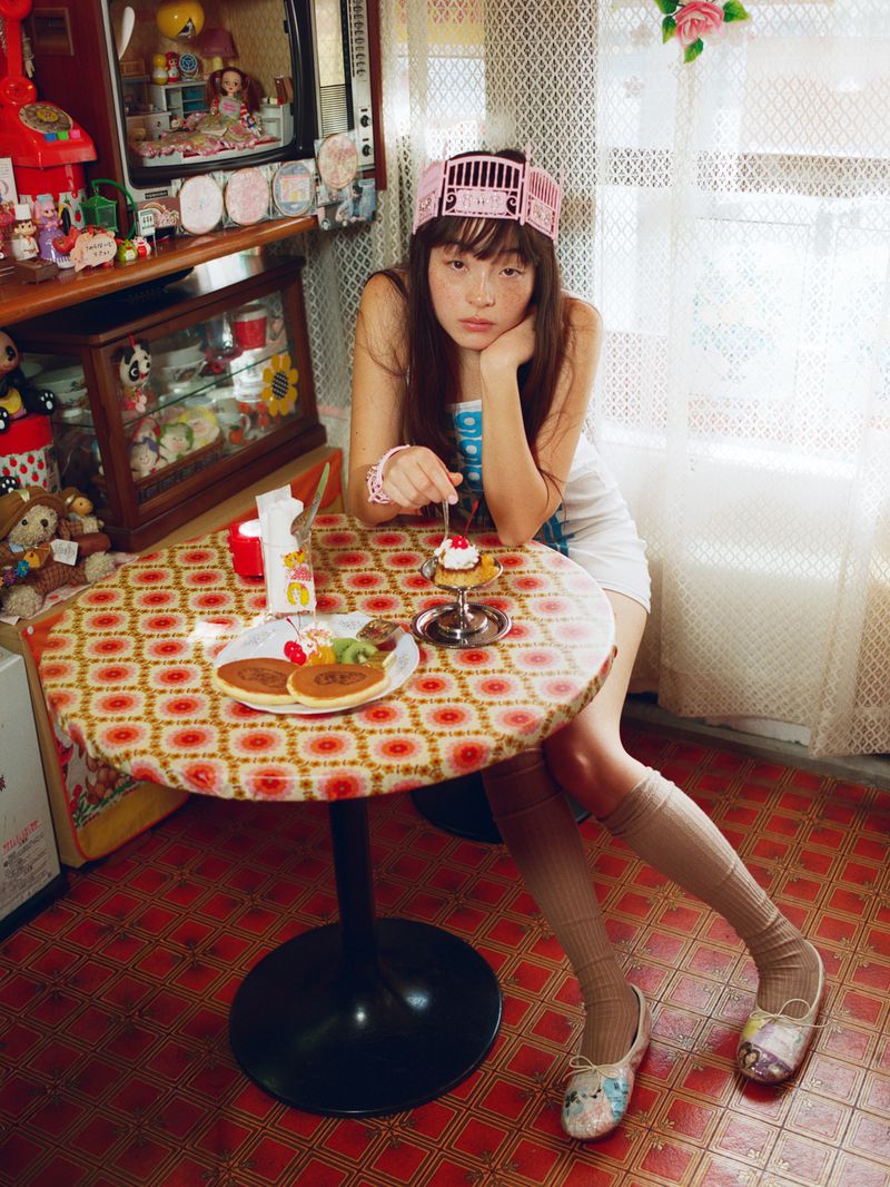 Serena Motola seated at a retro cafe table with pancakes and dessert, wearing a white mini dress and pink tiara headband.
