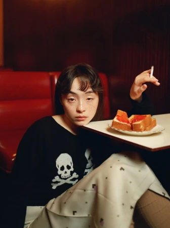 Serena Motola resting her head on a diner table beside a plate of jam toast while holding a cigarette in a red booth.