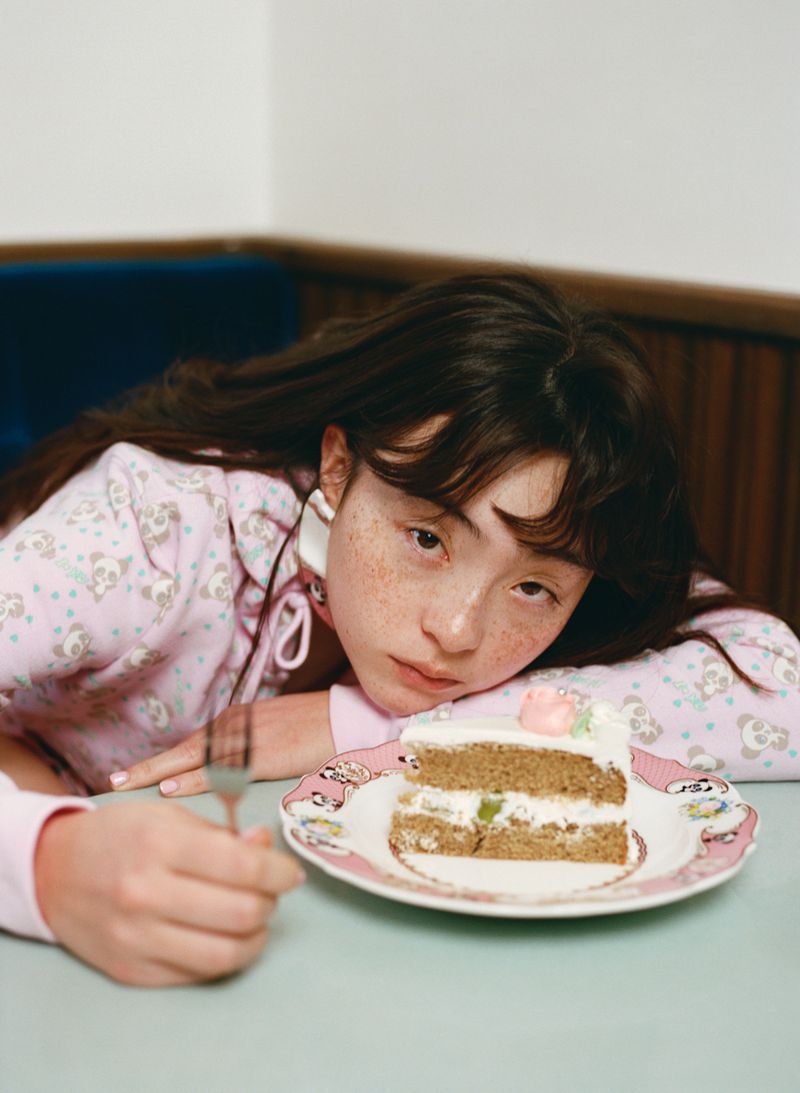 Woman in pink patterned top resting head on table beside a slice of cake on a plate for Ashley Williams SS26 Kissaten Diaries.