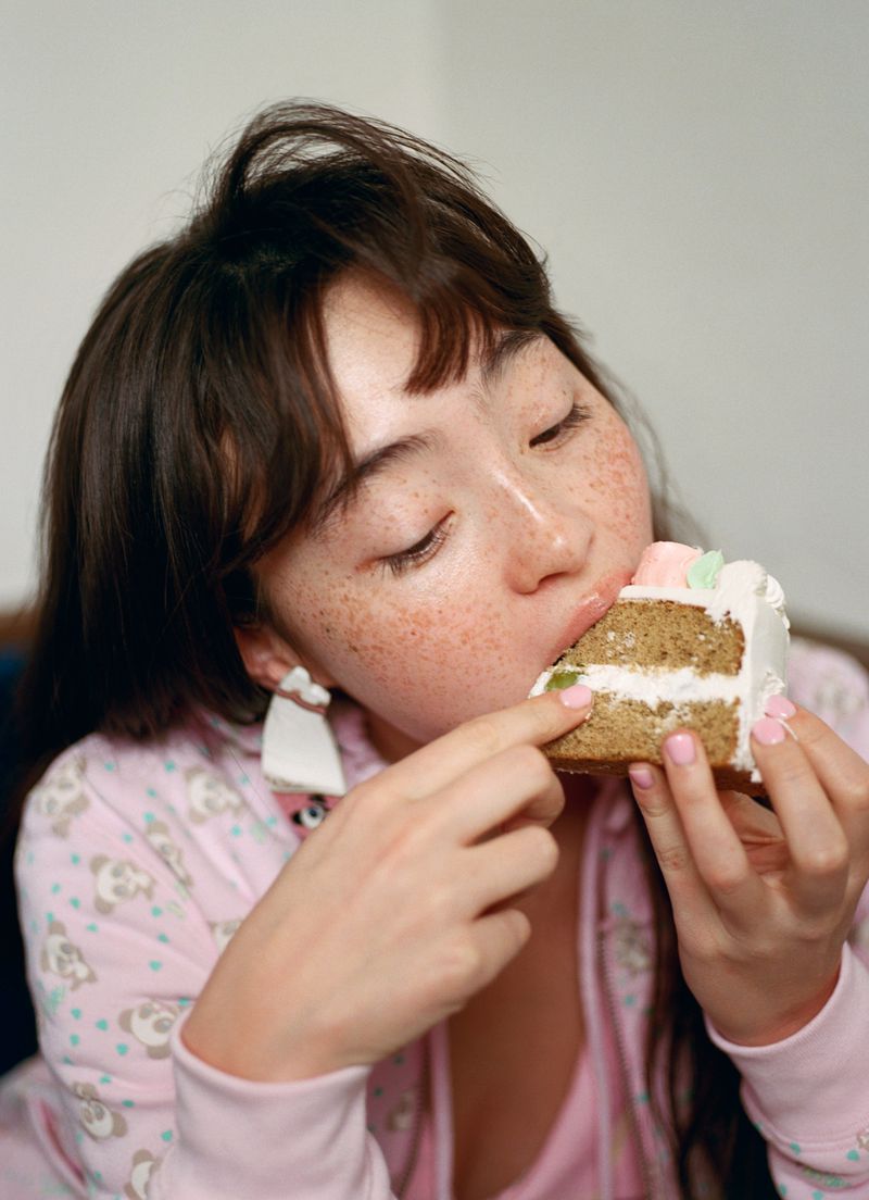 Close-up portrait of a young woman with freckles eating a slice of frosted layer cake while wearing pink patterned apparel