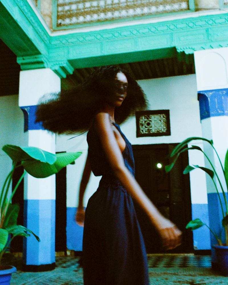 Fashion model in motion wearing a dark navy dress inside an architectural courtyard with blue pillars and potted plants.