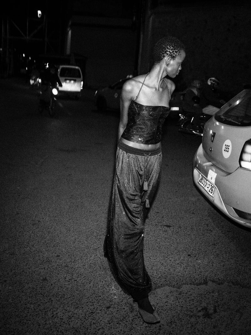 Model standing on a dark street at night next to a car, captured in high contrast monochrome photography.