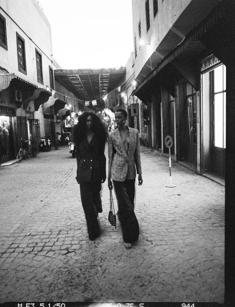 Two Black women in tailored blazers walk down a cobblestone alleyway in a black and white editorial shot.