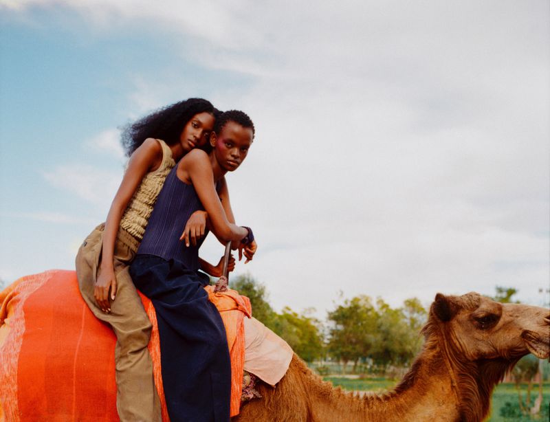 Two models seated together atop a camel wearing textured sleeveless tops and neutral trousers under an open daytime sky.