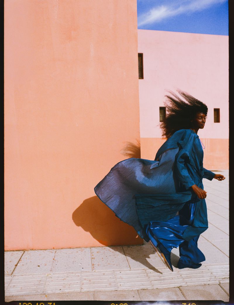 Model wearing a billowing blue robe walking in front of peach architectural walls with hair blowing in the wind.