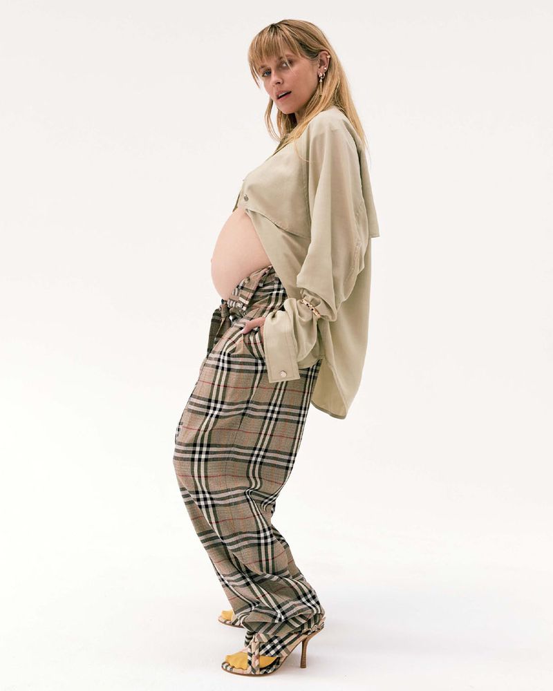 Pregnant woman poses in open beige shirt and plaid pants against a white background in a studio-style shot, looking at the camera.