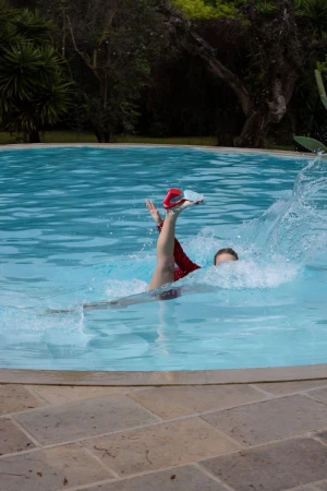 Model splashing into a bright blue swimming pool with one leg extended upward wearing a red high heel shoe.