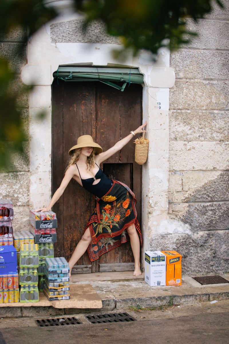 Model Manuela Frey posing in a wide-brim straw hat, black cut-out top, and printed wrap skirt in a rustic wooden doorway with drink crates.