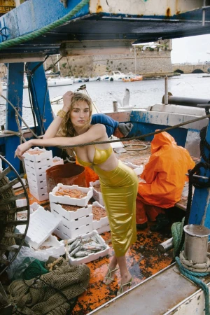 Model wearing a shiny yellow two-piece dress on a blue fishing boat surrounded by crates of fish and fishermen.